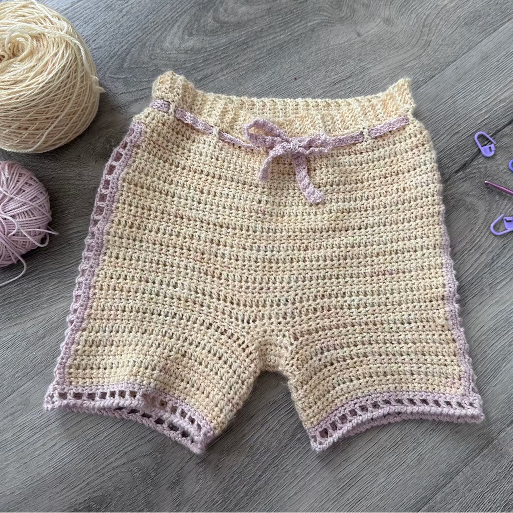 Crocheted shorts with pink trim on a wooden surface, accompanied by yarn and crochet tools.
