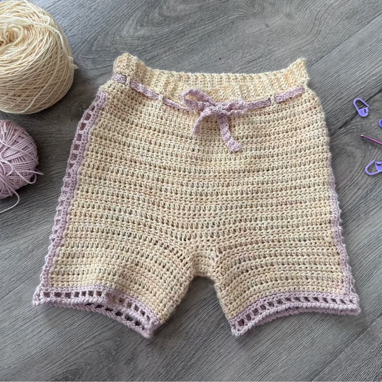 Crocheted shorts with pink trim on a wooden surface, accompanied by yarn and crochet tools.